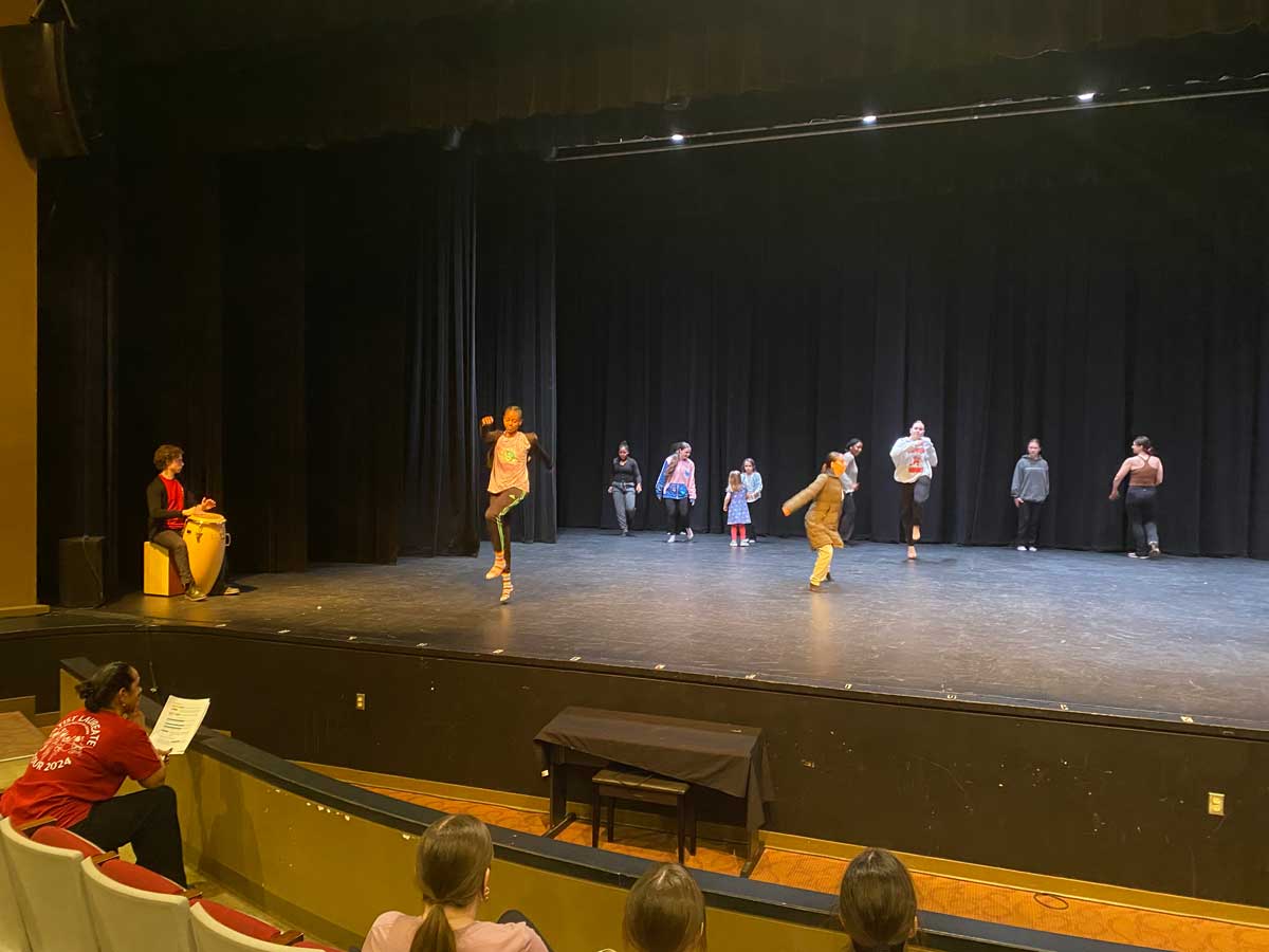 a student playing a drum while students practice a dance on stage while other students observe in the audience